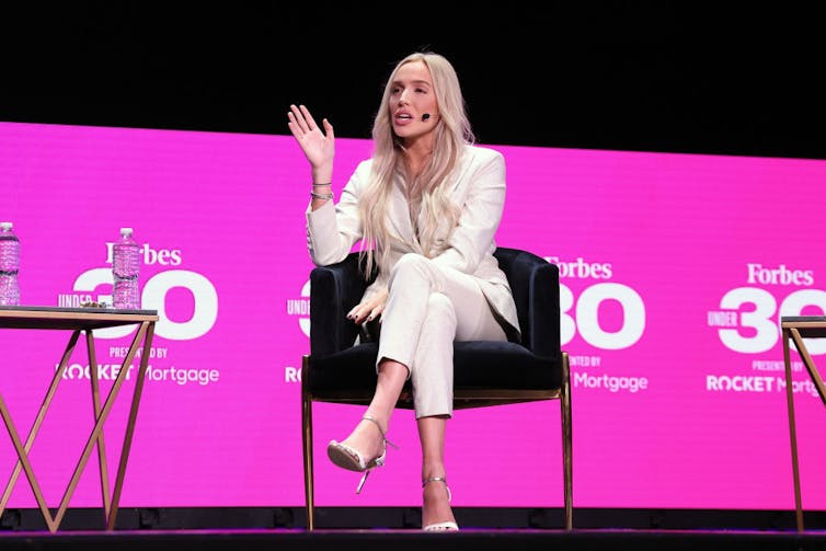 Woman sitting in chair speaks into microphone in front of pink backdrop.