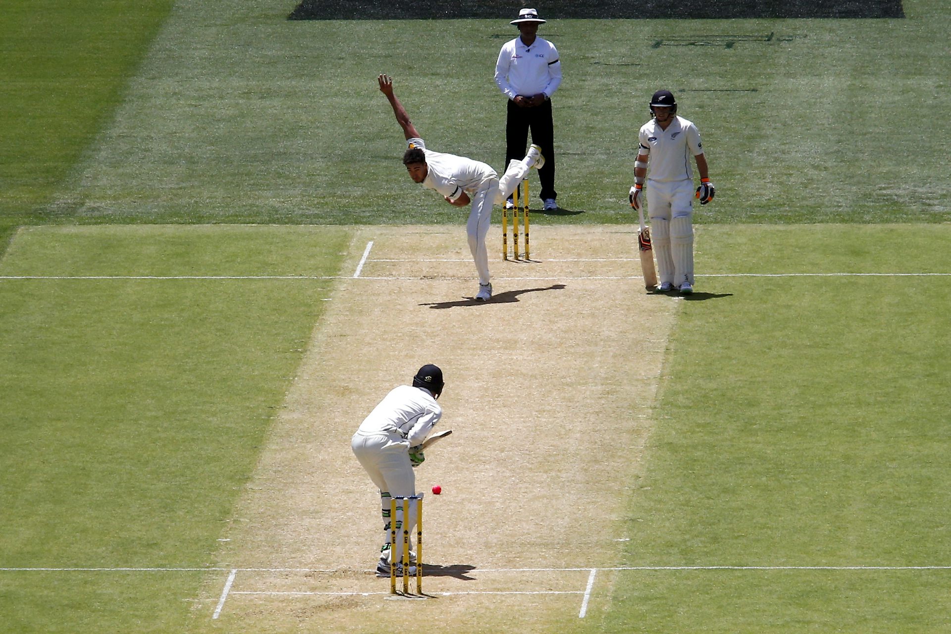 An Australian bowler bowls the ball to a New Zealand batter, while his partner looks on from the other end. All are wearing white uniforms.