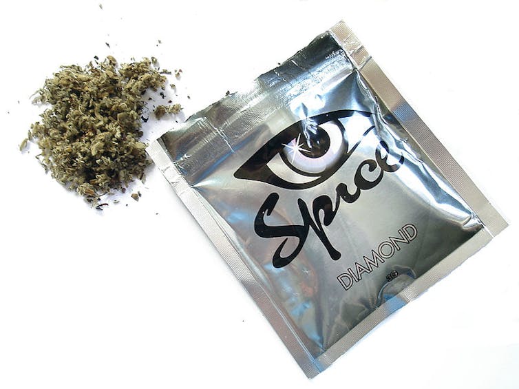 On a white background there is an opened packet of the drug 'Spice'. The wrapper is square, silver and says 'Spice' in black cursive lettering and 'Diamond' in embossed writing, There is an eye graphic on the wrapper.