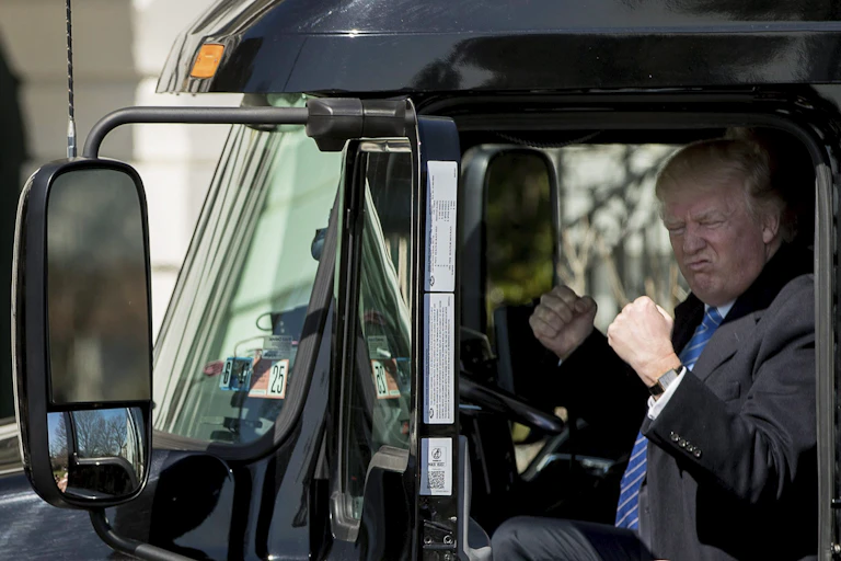 A man with greyish blond hair makes a face and raises his fists while in the cab of a truck.