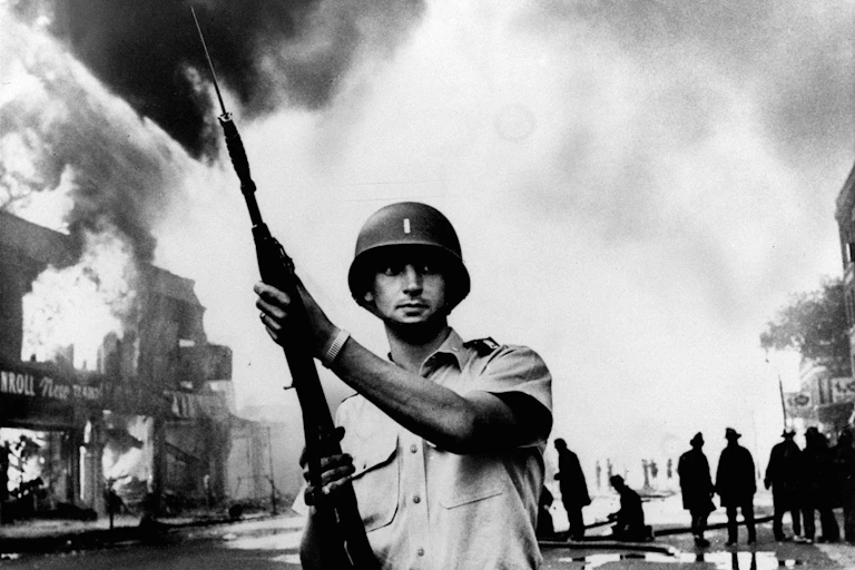 A white man wearing a uniform and a pith helmet holds a shotgun toward a threatening sky