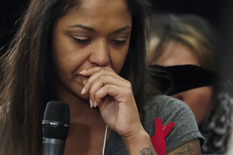 A dark-haired woman bows her head with tears on her face.
