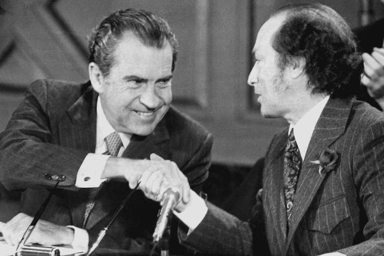 A man with thinning dark hair smiles at and shakes hands with another man with thinning dark hair.