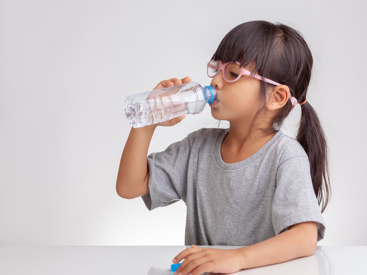 Niña con gafas rsas, flequillo y cole bebiendo agua de una botella de agua de plástico