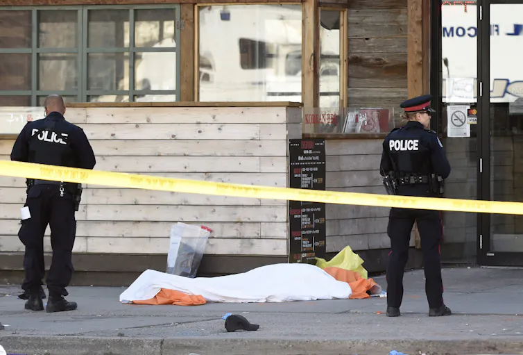 Police officers stand on a sidewalk behind yellow police tape near a body under a sheet