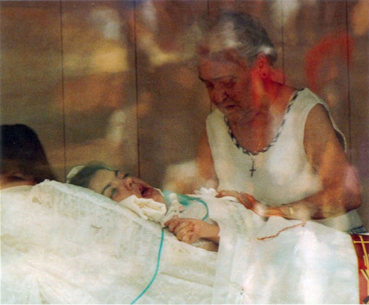 A girl with her mouth open lies in a bed as an older woman bends over her.