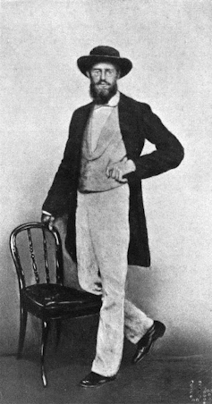 black and white photo of a standing man in a hat with one knee on a chair
