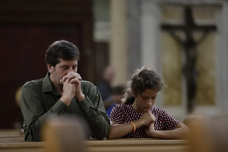 A man and a young girl seated in the pews with a crucifix in the background.