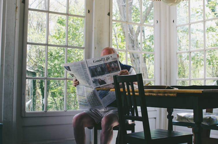 An older man sitting at a table in his house and reading a newspaper titled Good Life.