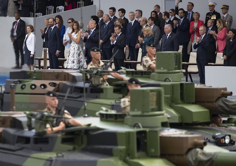 A row of green tanks go by on a street, with a small crowd of people watching from bleachers