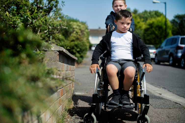 Boy in a wheelchair being assisted along a street by another boy