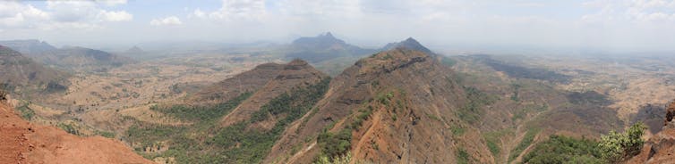 The ladscape of Maharashtra, India which is red in the left-hand corner of the photo and brown with dark green scrub as it progresses out to the surrounding hills and distance. The sky is slightly misty and blue with some clouds.