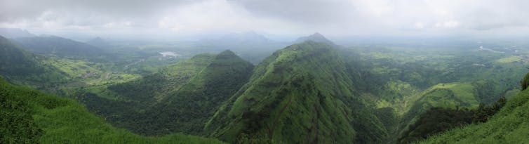 Photograph showing the same view as the previous image. This time the land is all lush greens and the mountainscape is clearer with a small lake in the distance. The sky is heavy with a light grey cloud before brightening up to a bolder white colour on the horizon.