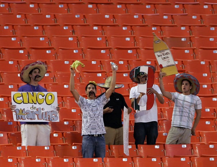 5 men wearing sombreros, mustaches and holding up signs in a baseball stadium. One sign says "Cinco de Marlins" and the other is a cut out of a beer bottle.