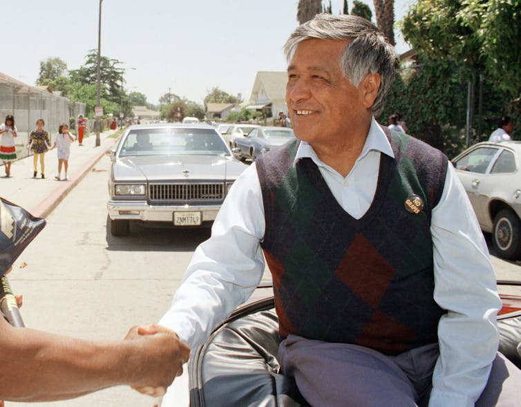 Photo of Cesar Chavez seated on back of car shaking hands in parade.