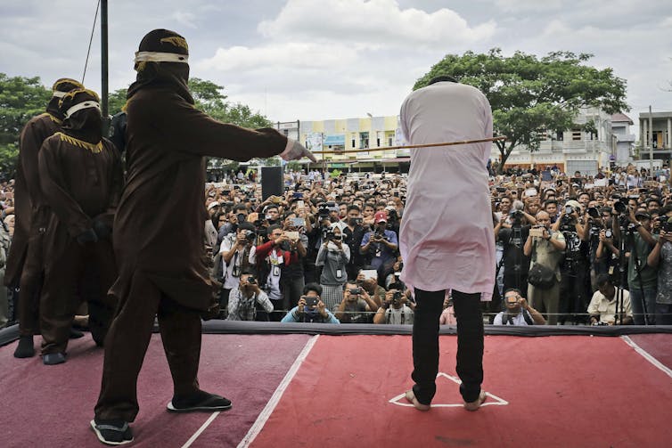Photo of man wearing hood caning another man. A large audience watches, with some holding cameras and phones, recording.