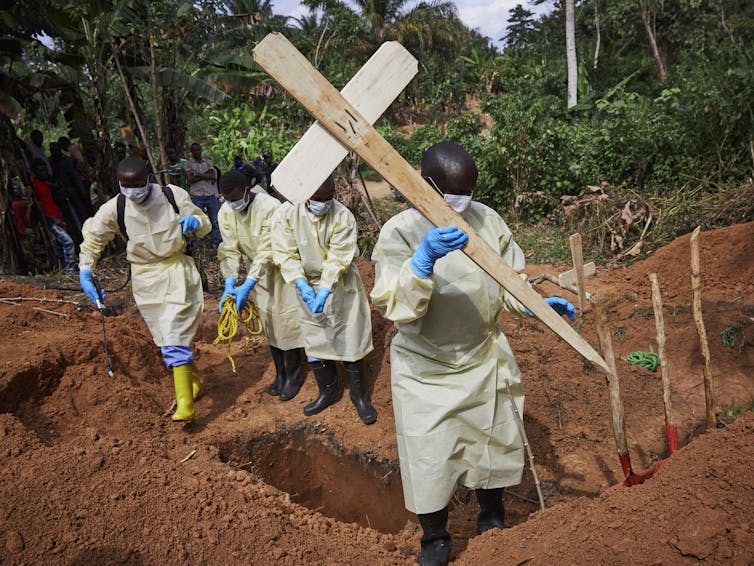 Four health workers in protective gear battling the outbreak unconventionally, one carries a crucifix, another disinfects a freshly dug grave. File 20190507 103057 yjf0iw.jpg?ixlib=rb 1.1