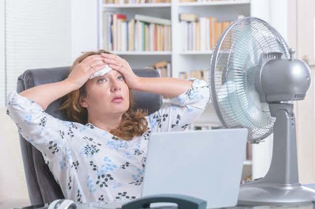 A woman sat at a desk with a large fan mops sweat from her brow.