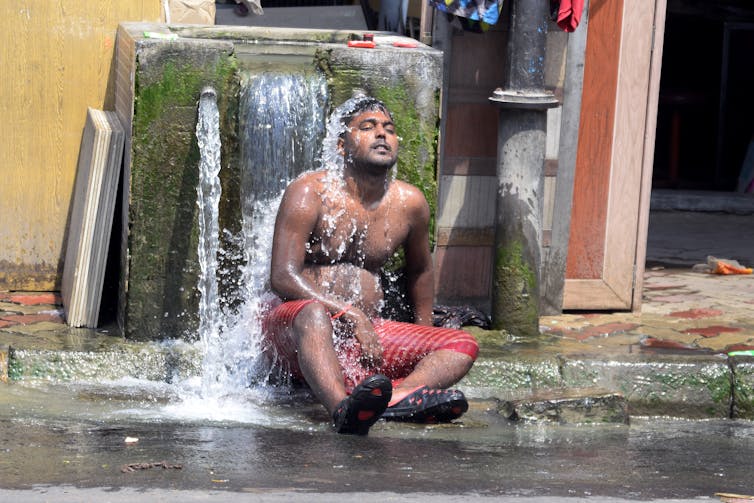 A shirtless man sits under a fountain to cool off.