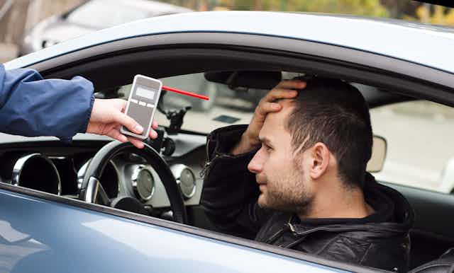 Person sitting in car with hand on forehead as another person extends a breathalyzer through their window.