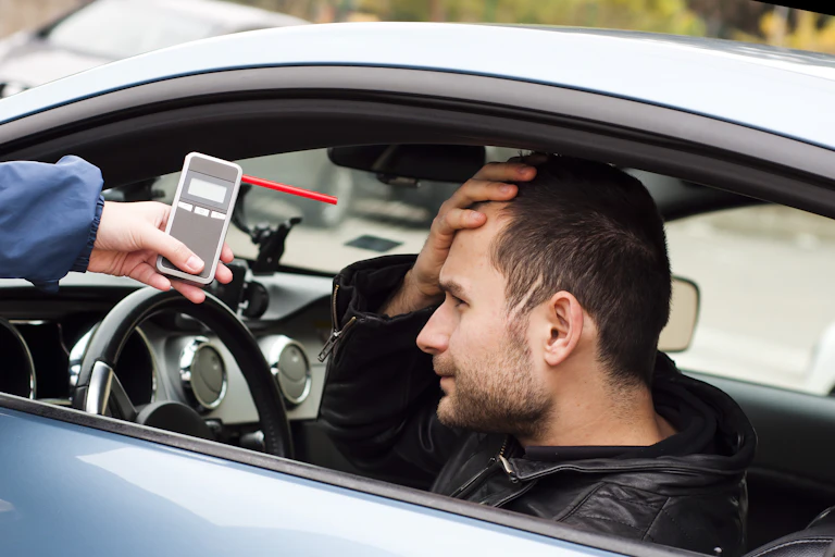 Person sitting in car with hand on forehead as another person extends a breathalyzer through their window.
