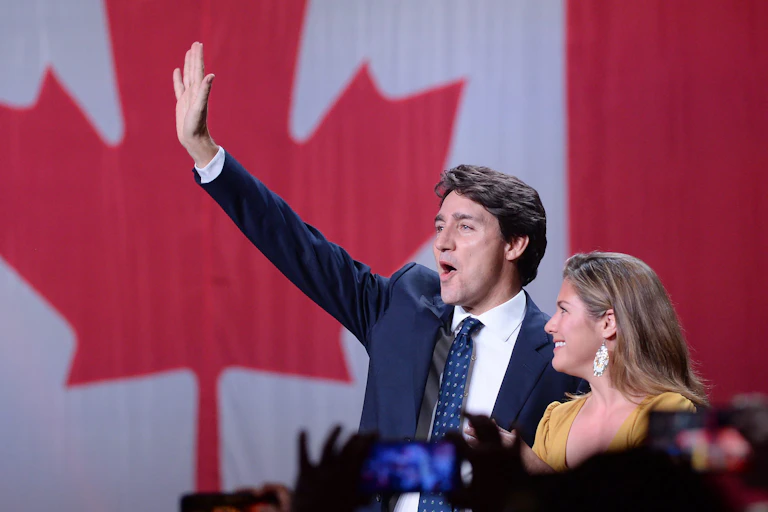 A dark-haired man waves while standing next to a woman with dark blonde hair. A large Canadian flag is behind him.