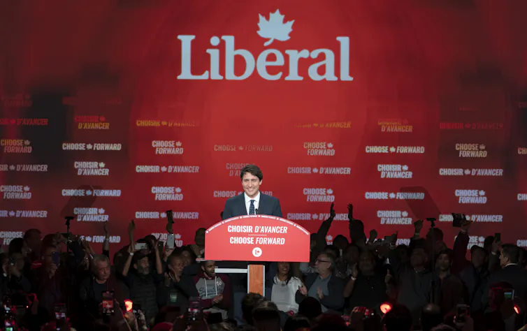 a man with dark hair smiles on a stage in front of a giant liberal party logo