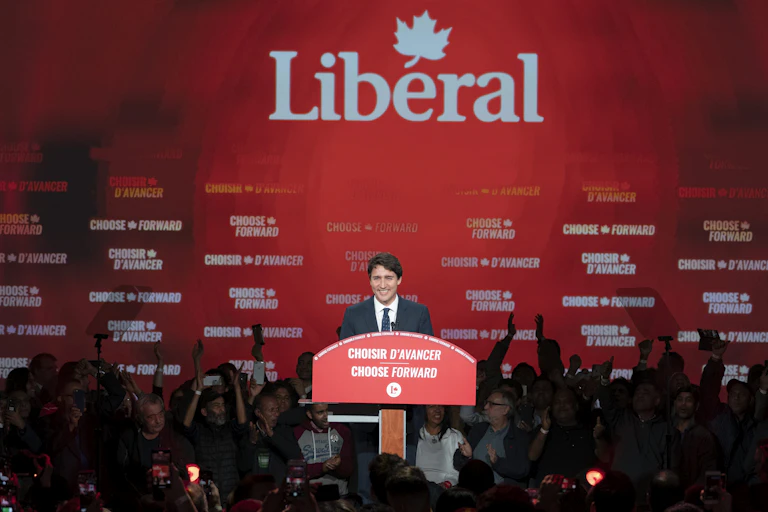 a man with dark hair smiles on a stage in front of a giant liberal party logo