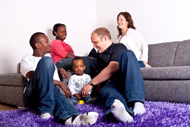 Four adults and a toddler sitting around a living room on couches and the rug, smiling.