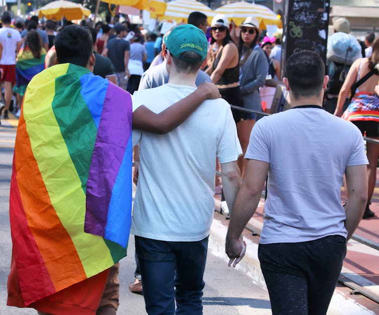 the backs of three people holding hands. One has a rainbow Pride flag draped over their shoulders