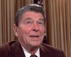 A smiling older man with brown hair in a brown suit.