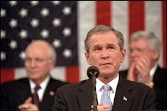 Photo of George Bush standing at microphone in front of American flag. Dick Cheney sits behind him to the left.