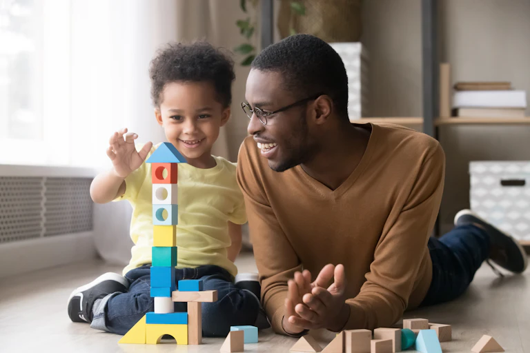 A Black child building a stack of blocks on the floor with his father