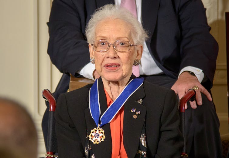 A photograph of Katherine Johnson seated wearing her newly awarded Presidential Medal of Freedom. She is sat in the centre of the photo with a man sat behind her. She wears a black blazer and orange blouse underneath, with small medals pinned into her top lapel. The medal hangs around her neck on a blue ribbon and shows a red and white star in the middle of a gold frame. In the very centre of the medal, there are white stars on a navy back ground. Katherine Johnson looks head on at the camera from behind her glasses and is poised semi-formally.