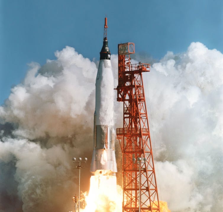 A rocket in the centre of the image takes off next to vertical red scaffold. Dark grey smoke engulfs the image from the left-hand corner to the top, becoming a white colour on the right-hand side. Bright white-yellow fire is showing from underneath the rocket as it lifts.