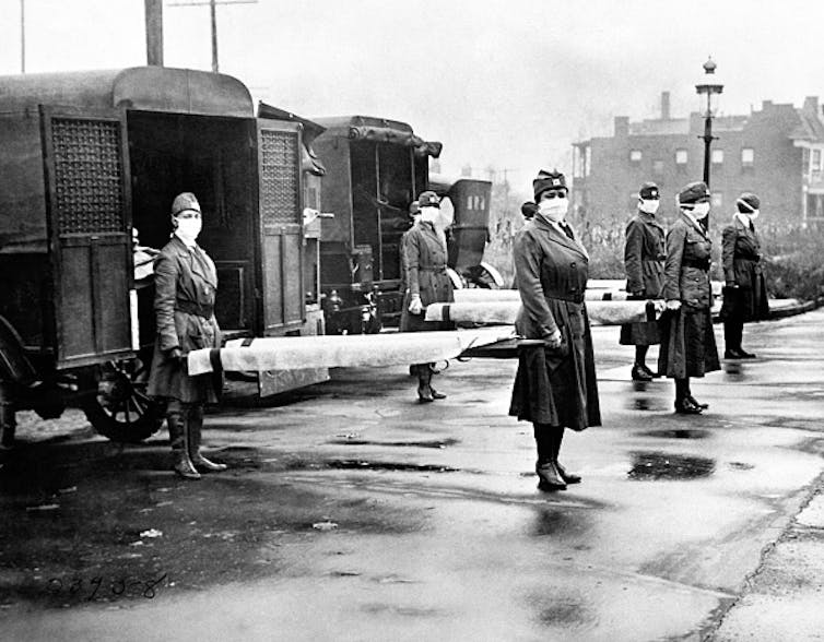 The St Louis Red Cross Motor Corps on duty with mask-wearing women holding stretchers at the backs of ambulances during the global flu epidemic