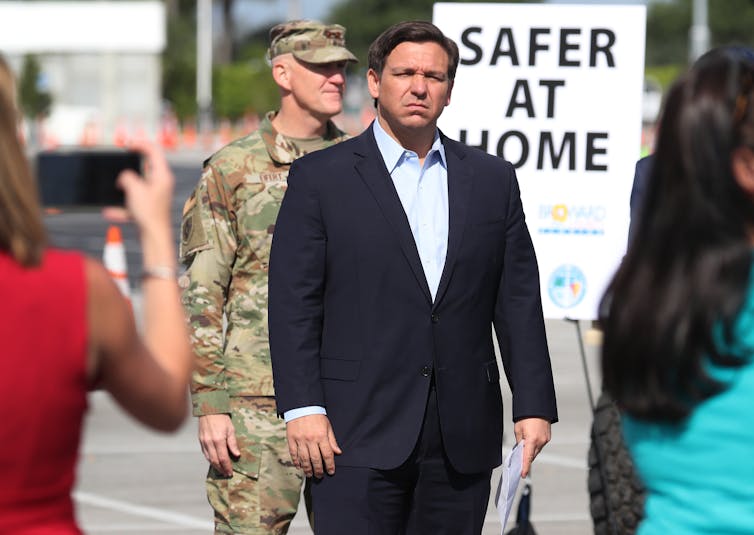 Gov. Ron DeSantis standing in front of Safer at Home sign