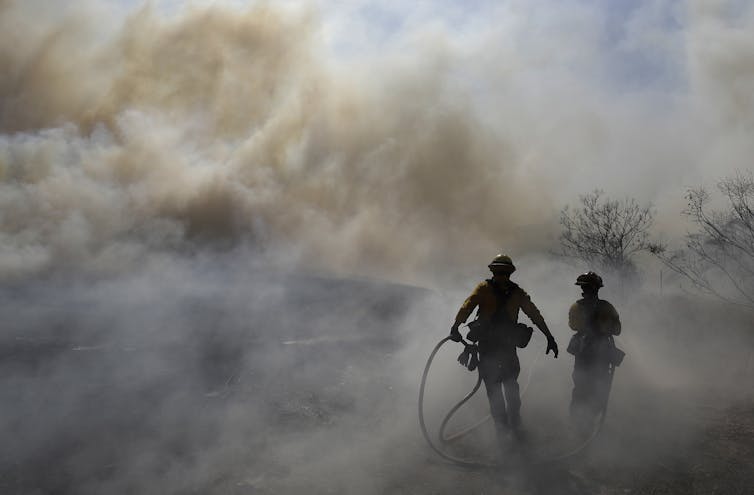 two firefighters carry equipment while walking towards smokey hillside, photo