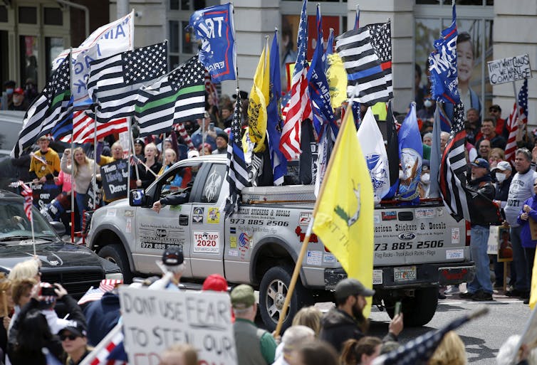 A photo of a city street with a parade of protesters waving flags that support first responders and former President Trump, and a silver pickup truck driving down the street in the center of the image with various flags and a "Trump 2020" sign on the side.