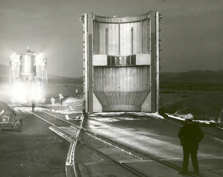 A black and white photo of a large, empty cylindrical structure, with a rocket releasing light in the background.