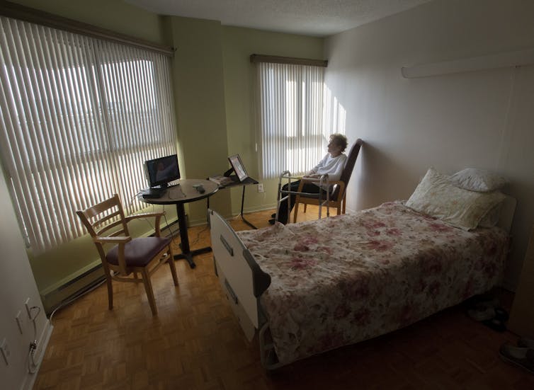 A woman sits in her room at a seniors' residence, blinds drawn.