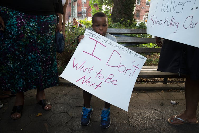 Small Black boy holding a sign that's nearly as big as him, saying "I don't want to be next" and in smaller letters "Stop gun violence, police brutality".