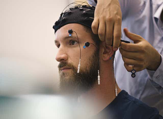 A technician's hands attach electrodes to a man being prepared for cognitive assessment with an electrode cap and electrodes on his face.