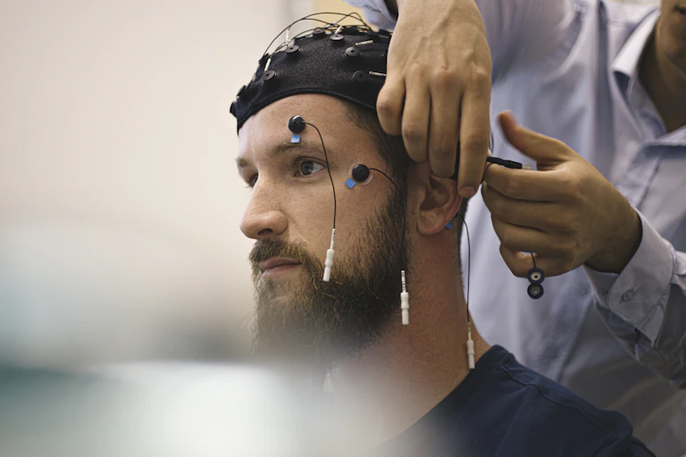 A technician's hands attach electrodes to a man being prepared for cognitive assessment with an electrode cap and electrodes on his face.