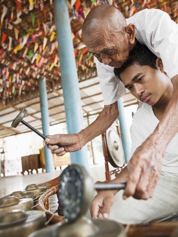 An older man leans over a younger man showing him a percussive instrument.