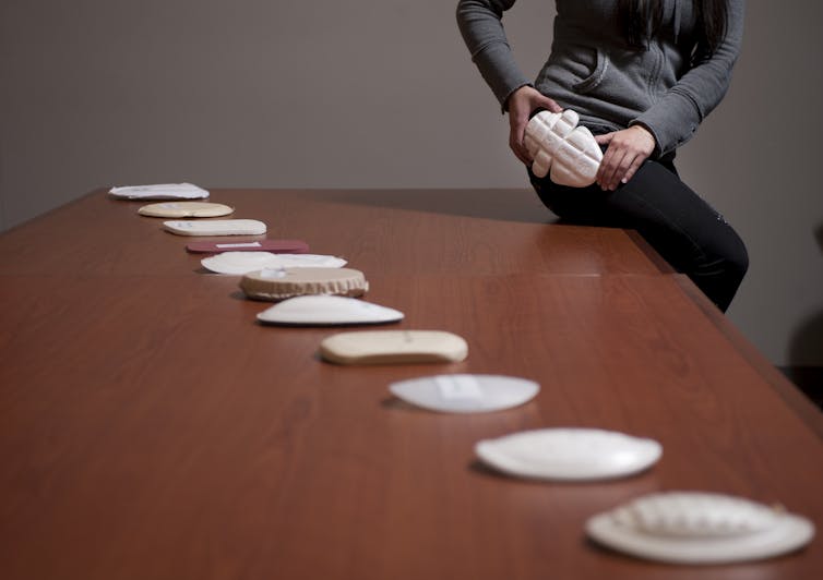 A woman sits on the corner of a table demonstrating the fit of a hip protector pad. Several types of hip pads are arranged on the table