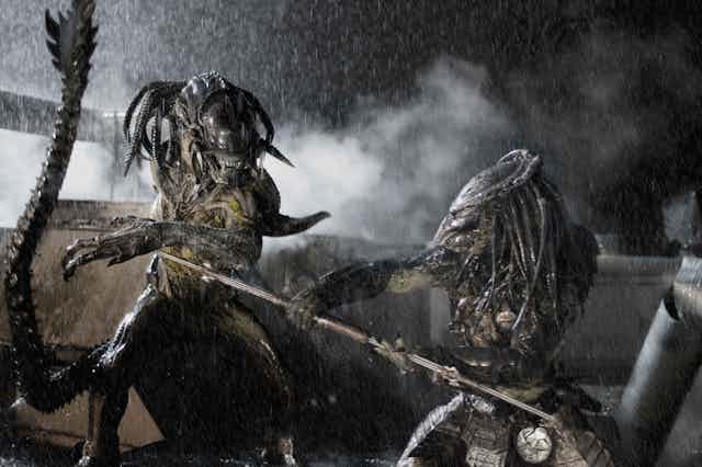 A scene from the move Alien vs. Predator shows two dreadlocked aliens fighting with spears in a rainstorm. 