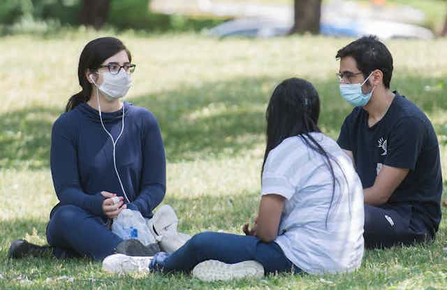 Young people sitting in a park wearing masks as they chat.