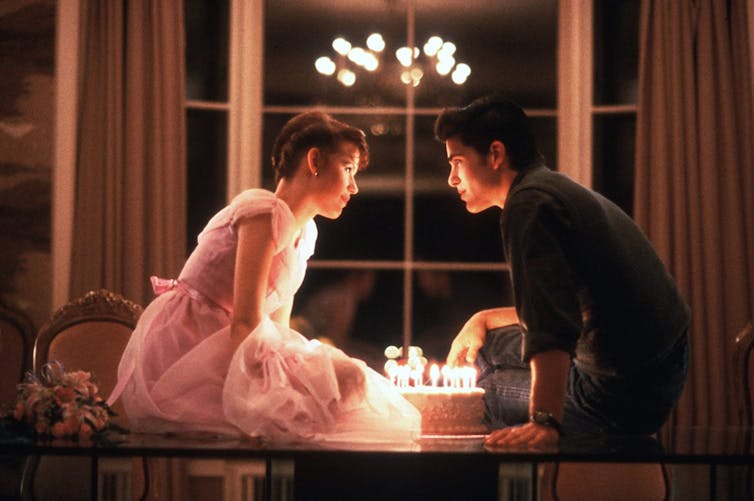 A man and a woman sitting on a table with a birthday cake filled with candles in between them