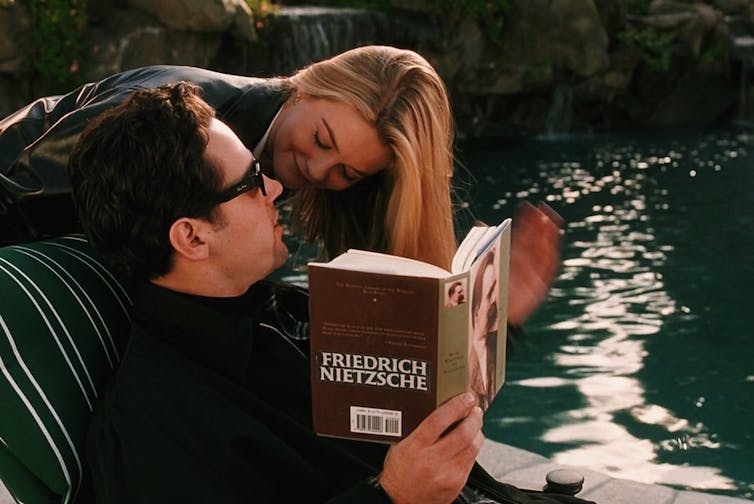 An image of a man sitting down reading a book and a woman leaning over his shoulder to look into his face